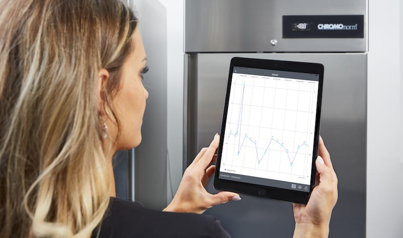 Digitale Temperaturüberwachung mit Tablet Eine Frau mit langen, blonden Haaren hält ein Tablet in den Händen und betrachtet eine graphische Auswertung mit Temperaturverläufen. Im Hintergrund steht ein Edelstahl-Kühlschrank von CHROMOnorm